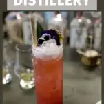 Red cocktail in tall glass with white foam and straw, purple flower garnish on gray bartop