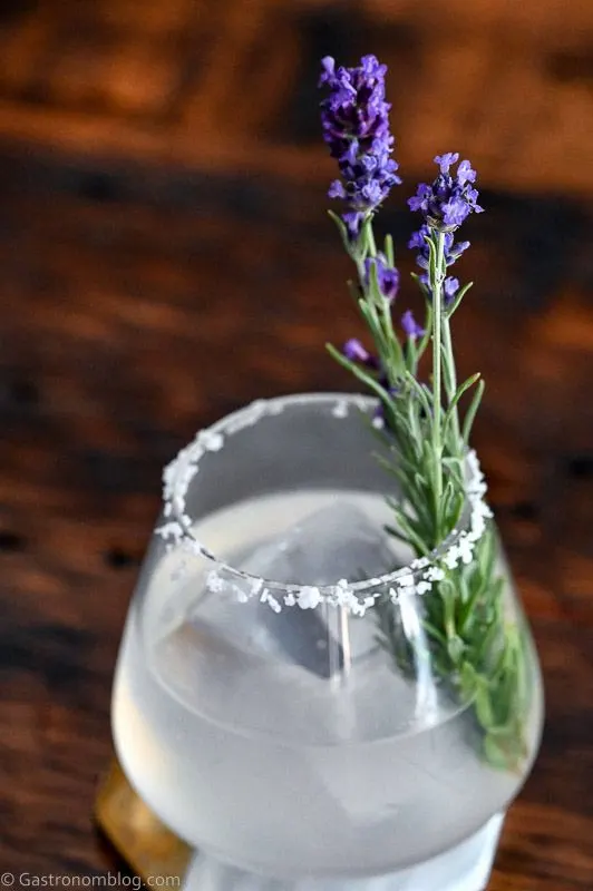 Opaque lavender margarita cocktail in rocks glass with salted rim, ice ball and lavender sprig on wooden table