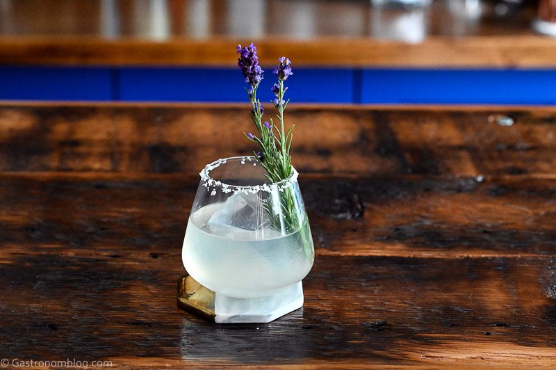 Opaque lavender margarita cocktail in rocks glass with salted rim, ice ball and lavender sprig on wooden table