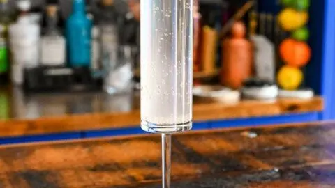 Hazy pink cocktail in tall glass on wood table, lilac garnish