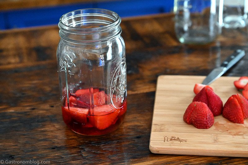 Cut strawberries covered with Campari in jar next to strawberries on cutting board