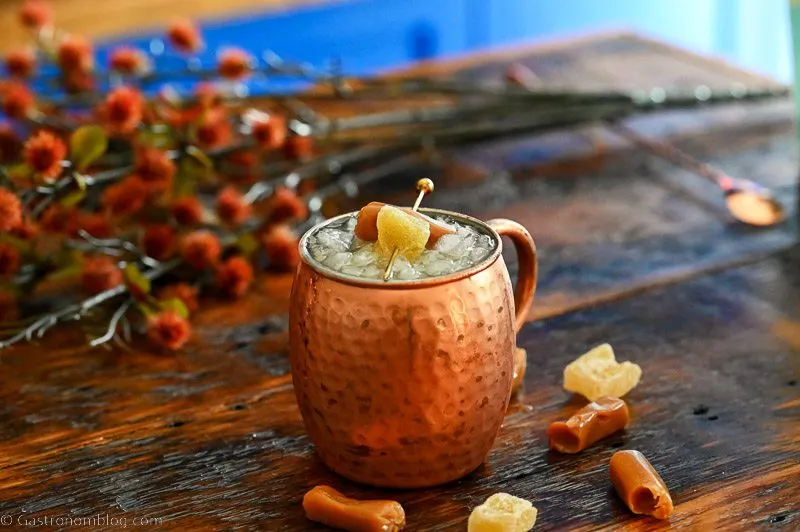 Copper Moscow mule mug with crushed ice and garnished with a caramel and candied ginger on a pick. Caramels and ginger in front of mug, orange flowers in background