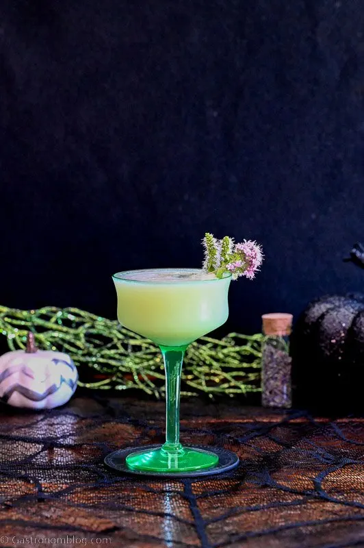 Green coupe with opaque cocktail with foam, mint flowers on side of coupe. Spider web napkin under and green glitter and pumpkins behind