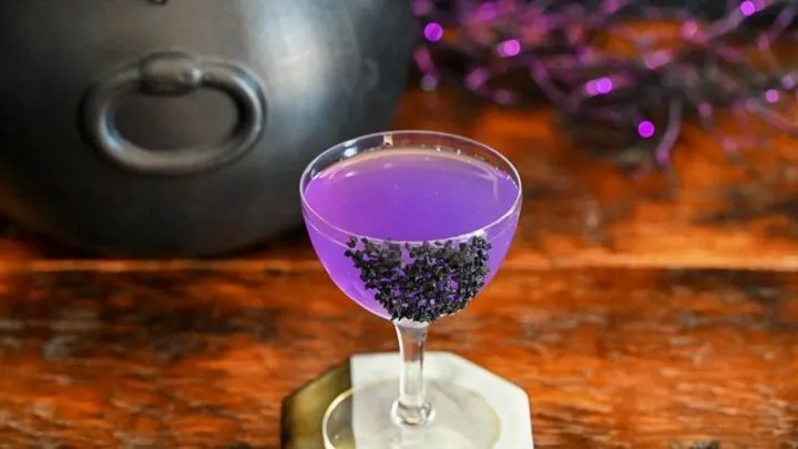 purple cocktail in coupe with black salt garnish, black cauldron and purple glitter sticks behind