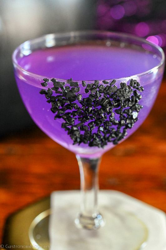 Sanderson Sisters Potion purple cocktail in coupe with black salt garnish, black cauldron and purple glitter sticks behind