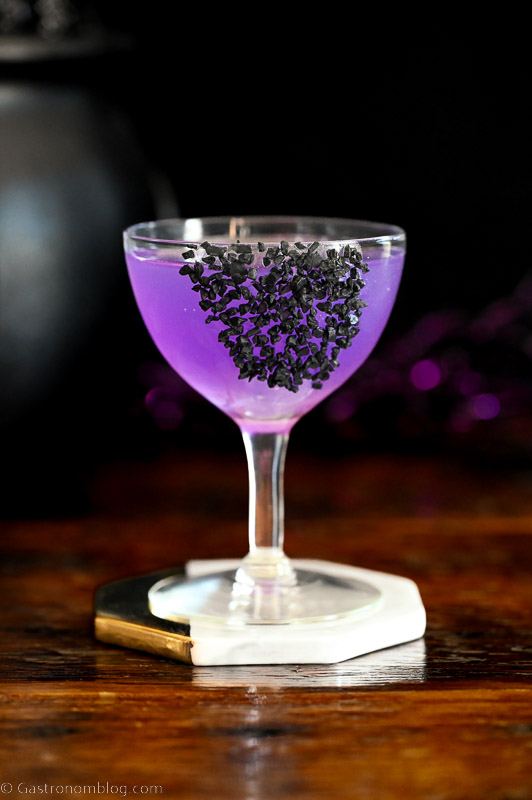 Sanderson Sisters Potion purple cocktail in coupe with black salt garnish, black cauldron and purple glitter sticks behind