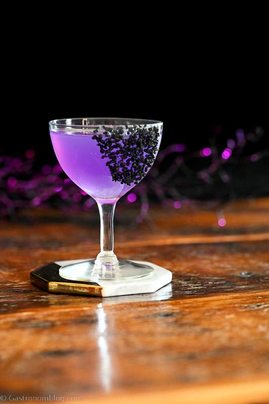 Sanderson Sisters Potion purple cocktail in coupe with black salt garnish, black cauldron and purple glitter sticks behind