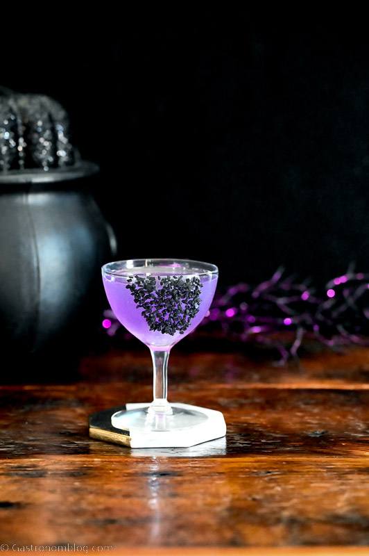 purple cocktail in coupe with black salt garnish, black cauldron and purple glitter sticks behind