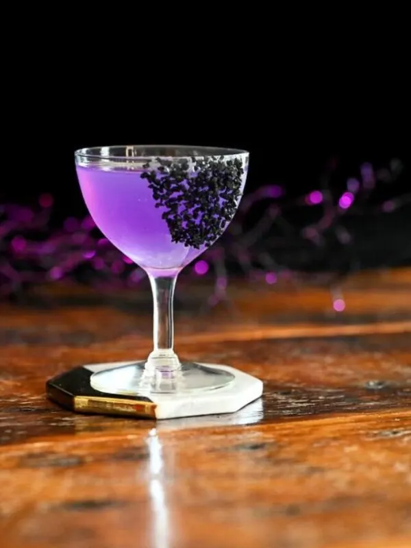 purple cocktail in coupe with black salt garnish, black cauldron and purple glitter sticks behind