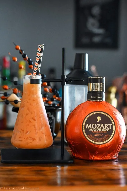 Orange Pumpkin Cocktail in a beaker as a glass with Halloween straws, pumpkin liqueur bottle next to glass