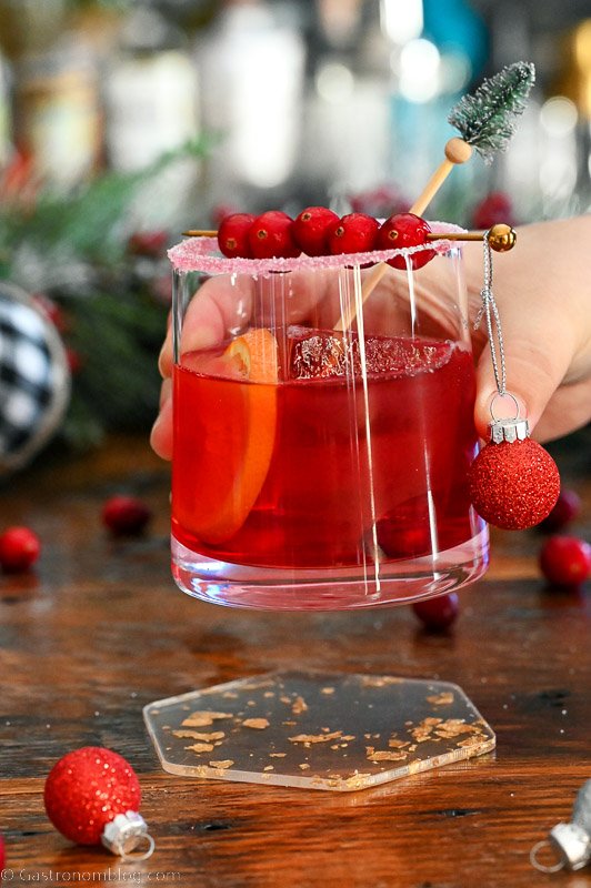 Christmas Old Fashioned red cocktail in rocks glass with orange slice, cranberries, pine stirrer and ornament.