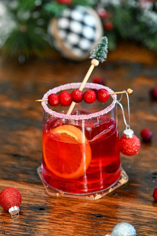 Christmas Old Fashioned red cocktail in rocks glass with orange slice, cranberries, pine stirrer and ornament.