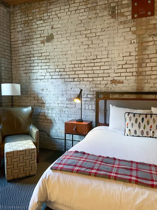 Bed in hotel room at Hewing Hotel, Minneapolis. Plaid bedspread, white sheets, nightstand with lamp