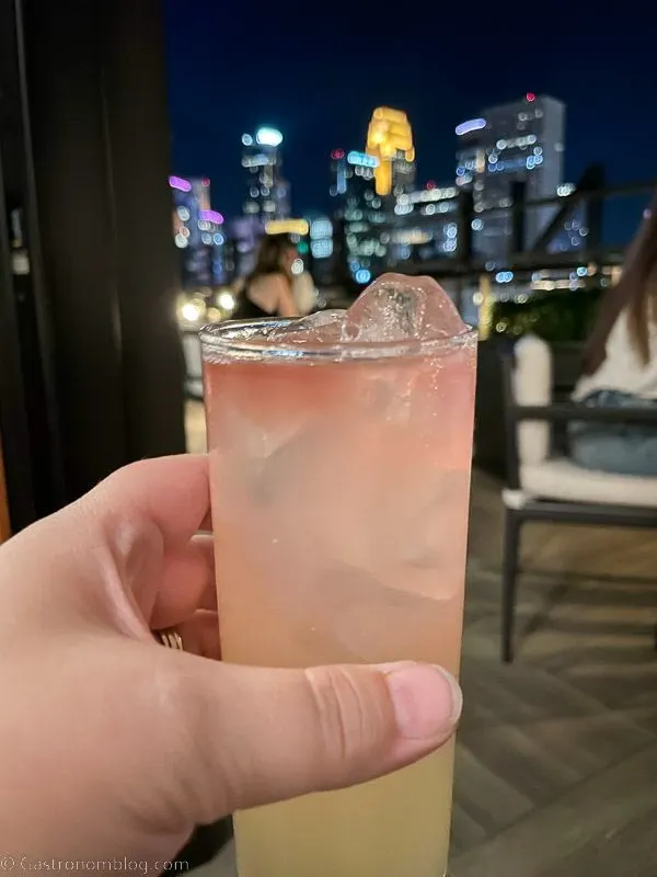 Hand holidng light pink cocktail in glass up against Minneapolis skyline at night