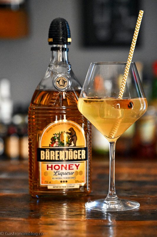 Yellow Thanksgiving Champagne Cocktail in a wine glass with straw. Barenjager honey liqueur bottle next to glass.