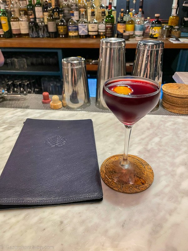 dark red cocktail in coupe with orange peel and leather menu on white marble bar