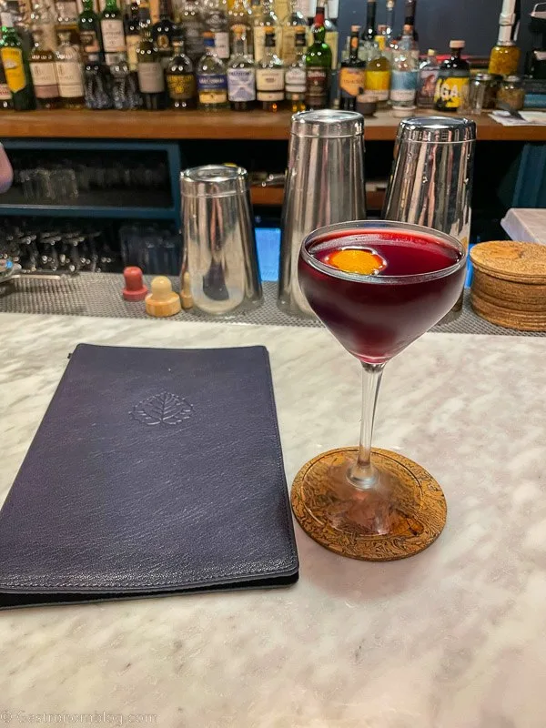 dark red cocktail in coupe with orange peel and leather menu on white marble bar