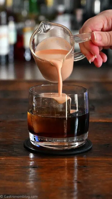 orange cream being poured into brown cocktail in rocks glass