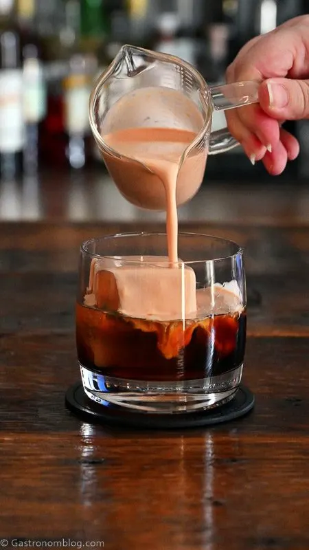 orange cream being poured into brown cocktail in rocks glass