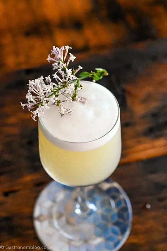 Yellow cocktail in glass with white foam top. Lilac sprig garnish on Lilac Gin Sour Cocktail.