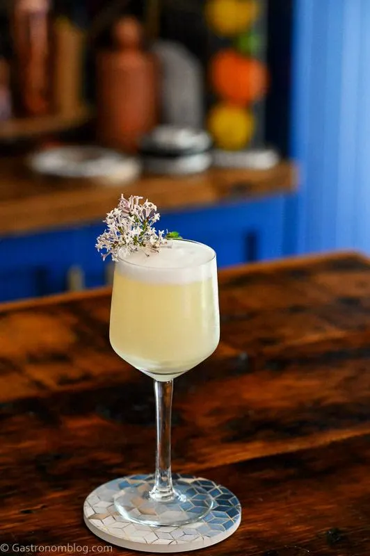 Yellow cocktail in glass with white foam top. Lilac sprig garnish on Lilac Gin Sour Cocktail.