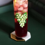 Purple grape lemonade cocktail in tall glass with green grapes print on green table