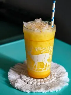 Yellow citrus fizz cocktail in a glass with a deer print on it. On a white coaster, on a blue table.