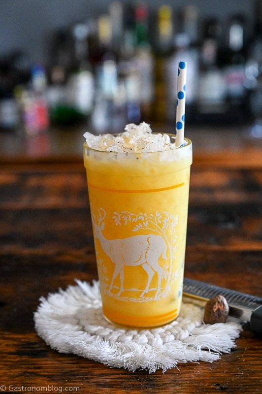 Yellow citrus fizz cocktail in a glass with a deer print on it. On a white coaster, on a wood table.