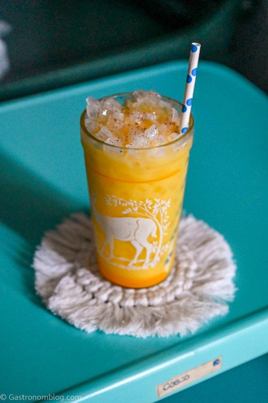 Yellow citrus fizz cocktail in a glass with a deer print on it. On a white coaster, on a blue table.