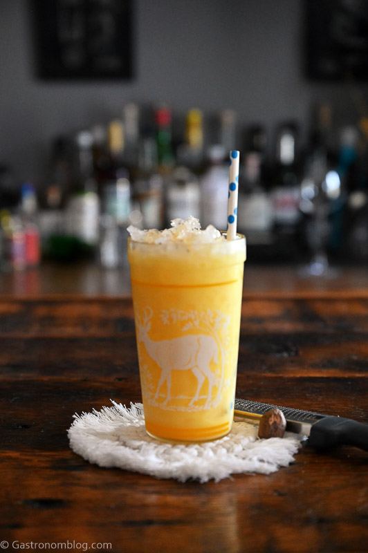 Yellow citrus fizz cocktail in a glass with a deer print on it. On a white coaster, on a wood table.