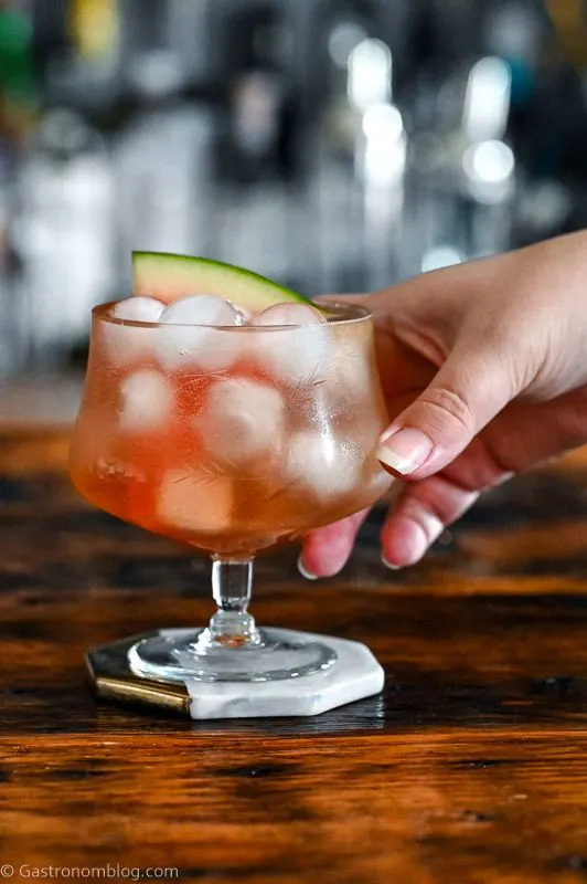 Pink cocktail, Watermelon Aperol Spritz in a low glass with stem on wood table. Ice cubes in glass with watermelon slice, hand holding