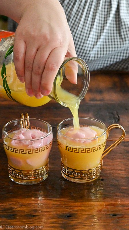orange juice being poured into gold rimmed punch cup filled with raspberry sherbet