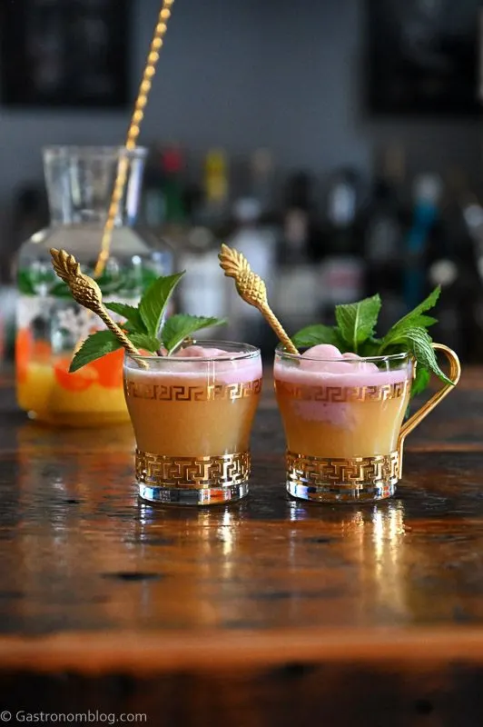 orange pineapple orange punch in gold trimmed punch cups topped with raspberry sherbet and mint leaves, pitcher with oranges print behind