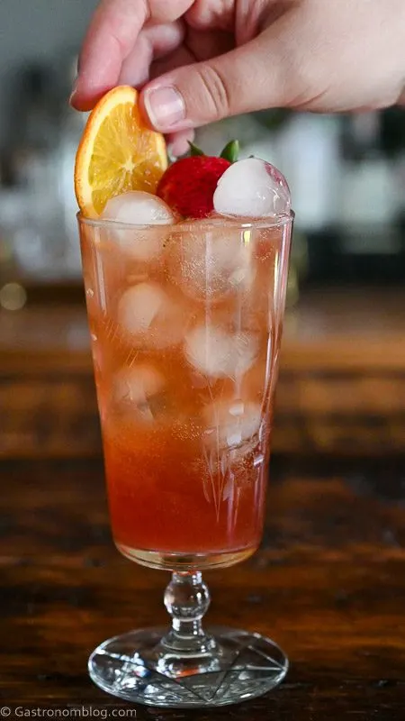 pink Strawberry Aperol Spritz cocktail with ice balls, orange slice and strawberry in tall glass on wood table