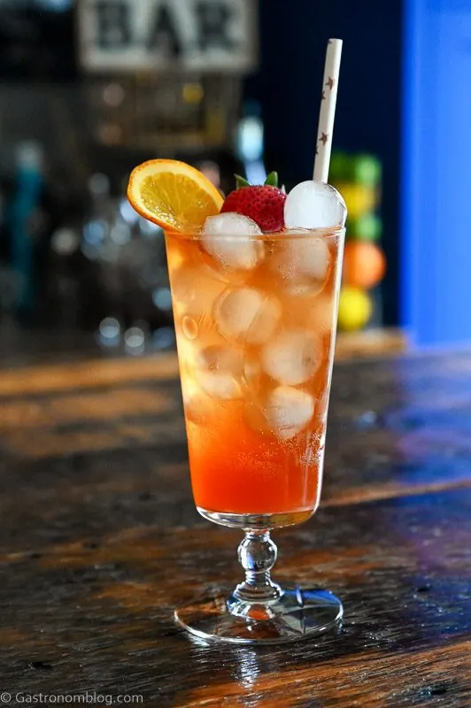 pink Strawberry Aperol Spritz cocktail with ice balls, orange slice and strawberry in tall glass on wood table