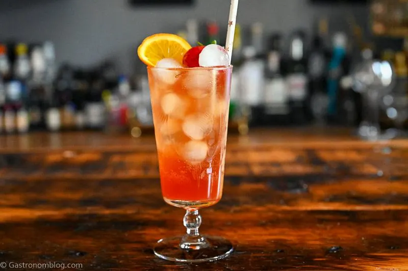 pink Strawberry Aperol Spritz cocktail with ice balls, orange slice and strawberry in tall glass on wood table