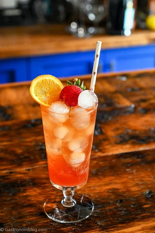 pink Strawberry Aperol Spritz cocktail with ice balls, orange slice and strawberry in tall glass on wood table