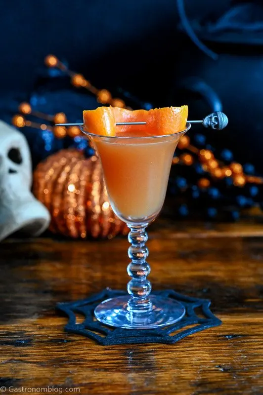 orange Halloween cocktail recipe in coupe with orange peel and Halloween decor behind