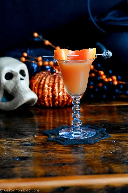 orange Halloween cocktail recipe in coupe with orange peel and Halloween decor behind