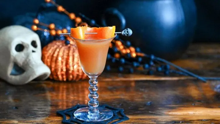 orange Halloween cocktail recipe in coupe with orange peel and Halloween decor behind