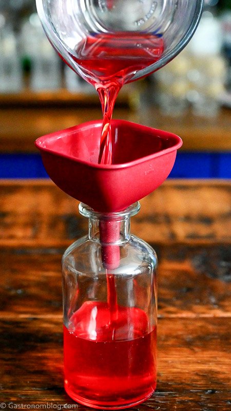 pouring pink cranberry simple syrup through red funnel into glass bottle