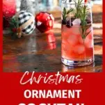 red ornament cocktail in tall glass with ice, rosemary sprigs and cranberries. ornaments and tree bottle brushes behind