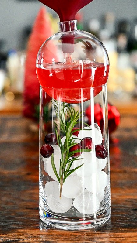 red ornament cocktail in tall glass with ice, rosemary sprigs and cranberries. ornaments and bottle brushes behind