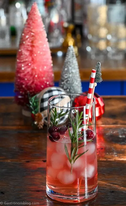 red ornament cocktail in tall glass with ice, rosemary sprigs and cranberries. ornaments and bottle brushes behind