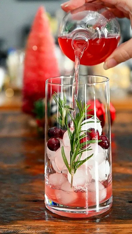 red ornament cocktail in tall glass with ice, rosemary sprigs and cranberries. ornaments and bottle brushes behind