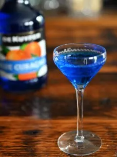 So what is blue curacao? This blue liqueur is in a cocktail coupe, and in a bottle behind on a wood table