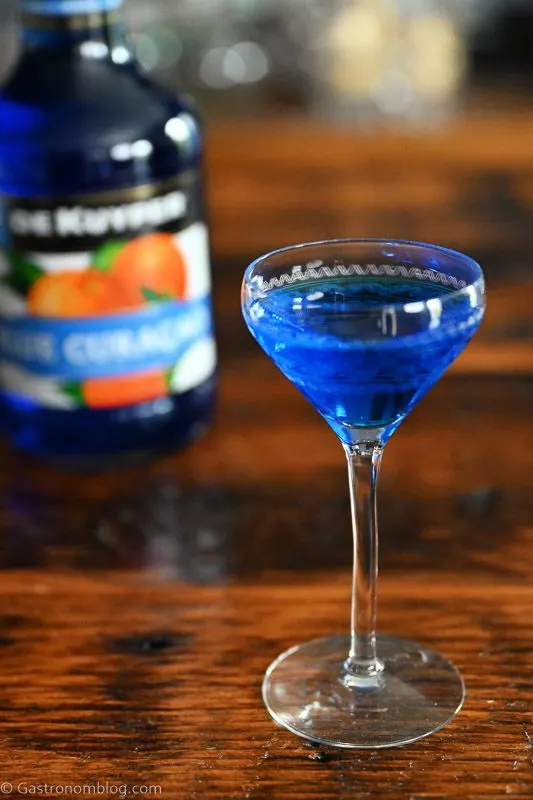 So what is blue curacao? This blue liqueur is in a cocktail coupe, and in a bottle behind on a wood table