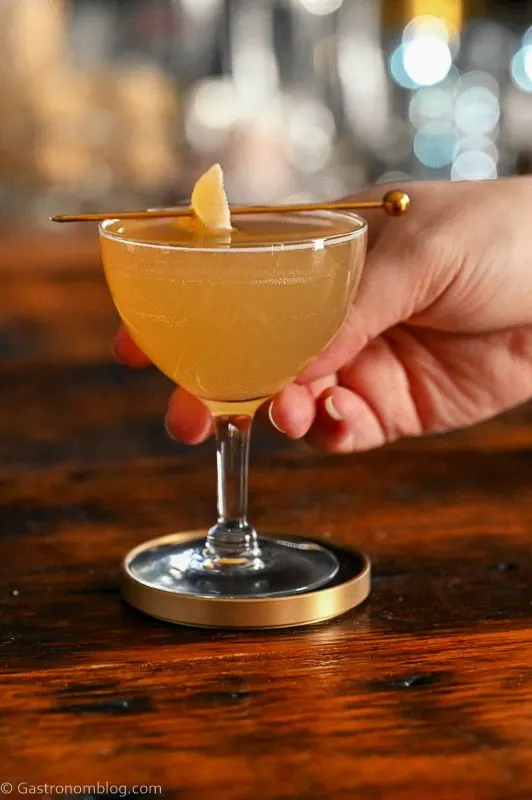 gold colored maple mocktail in cocktail coupe with crystallized ginger garnish on cocktail pick. On wood table.