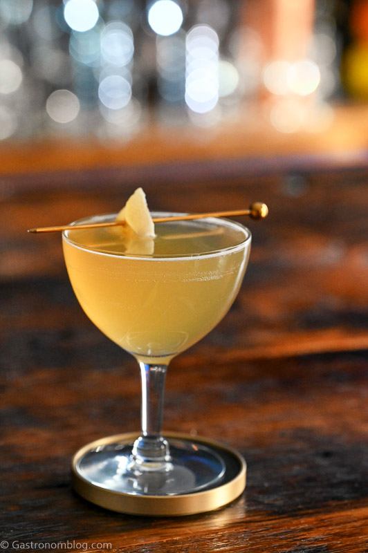 gold colored maple mocktail in cocktail coupe with crystallized ginger garnish on cocktail pick. On wood table.