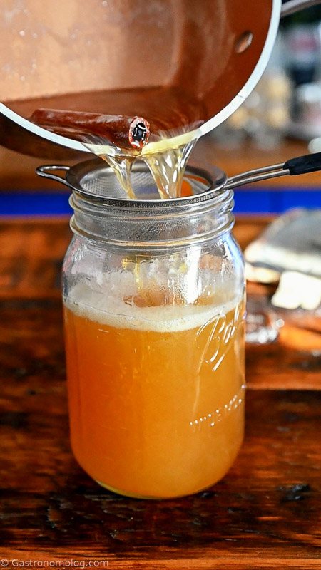 straining spiced honey syrup through a fine mesh strainer into large mason jar